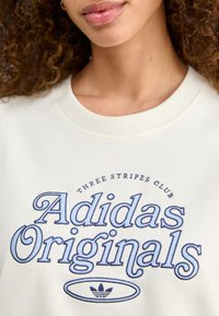 Vit sweatshirt med blå broderad text "Adidas Originals" och "Three Stripes Club", med rund halsringning och mjuk textur.