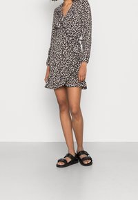 Kobieta w długim rękawie w sukience o wzorze lamparta z falbanami, w zestawieniu z czarnymi sandałami na otwartym palcu na jednolitym białym tle.