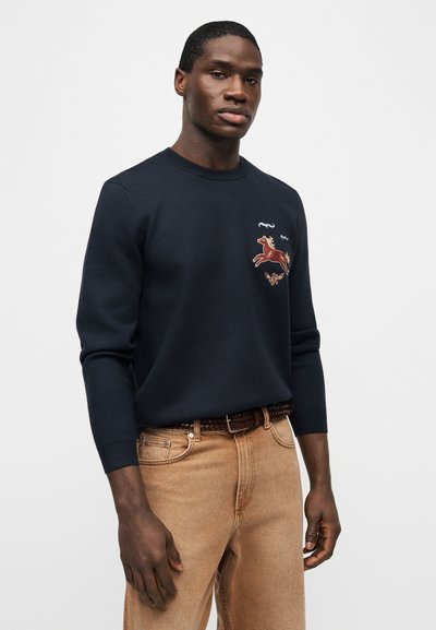 Άνδρας που φοράει μαύρο φούτερ με κεντημένο άλογο και πεταλούδα, μπεζ τζιν με πλεκτή καφέ ζώνη, στέκεται μπροστά σε ένα απλό φόντο.