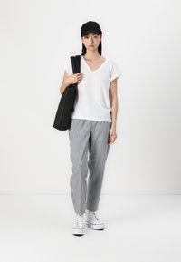 Vit kortärmad T-shirt med V-hals, grårandiga byxor, vita sneakers och en svart keps; modell som håller en svart väska.