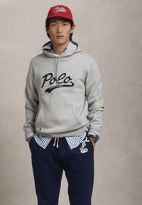 Felpa grigia in cotone con logo "Polo", indossata sopra una camicia a righe blu e bianche. Pantaloni della tuta blu con un emblema, indossati con un cappellino rosso.