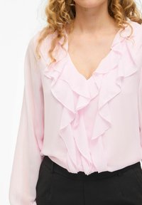 Gaiši rozā šifona blūze ar ruffles apkakli un garām piedurknēm, apvienota ar melniem biksēm. Audums izskatās maigs un plūstošs.