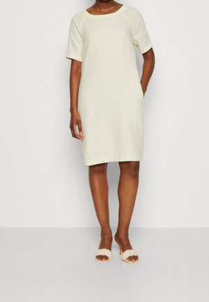 Kvinde iført en knælængde, kortærmet, cremehvid kjole med lommer og matchende flettet strop-sandaler med høje hæle.