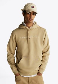 Beżowa bluza z kapturem z nadrukowanym logo "TOMMY JEANS", regulowanym sznurkiem i przednią kieszenią; zestawiona z beżową czapką z naszywką z logo.