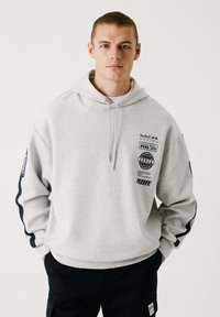 Sudadera gris claro con gráficos en el frente y las mangas, con capucha ajustable. Mangas largas con acentos en azul oscuro. Textura suave, ajuste casual.