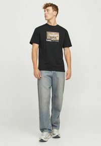 Sort bomulds t-shirt med et grafisk design med teksten "Later", parret med lysblå denimjeans og hvide sneakers.
