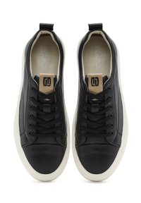 Sneakers in pelle nera con profilo basso, suola in gomma bianca, lacci neri e una toppa con logo marrone sulla linguetta. Superficie liscia, design casual.