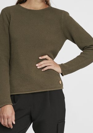 Kobieta ubrana w oliwkowo-zielony teksturowany sweter z długim rękawem i czarne spodnie, z ręką opartą na biodrze na jednolitym tle.