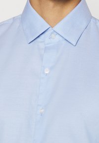 Lichtblauw overhemdd met een gestructureerde stof, puntkraag en witte plastic knopen. Bevat een klein logodetail op de borst.