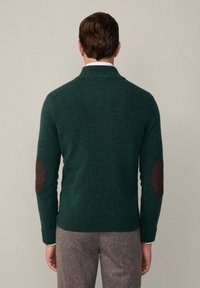 Grüner Wollpullover mit Schalkragen und braunen Ellenbogenpatches. Strukturstrick, figurbetonter Schnitt und gerippte Bündchen und Saum.