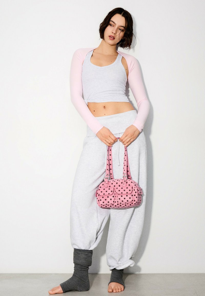 Femme en débardeur gris clair et pantalon avec manches longues roses tient un sac à main rose à pois, debout pieds nus contre un mur blanc.