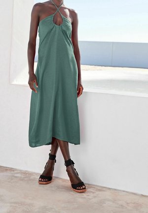 Groene mouwloze jurk met een keyhole halslijn en geplooide details. Gecombineerd met zwarte platform sandalen met enkelbandjes. Soepele textuur.