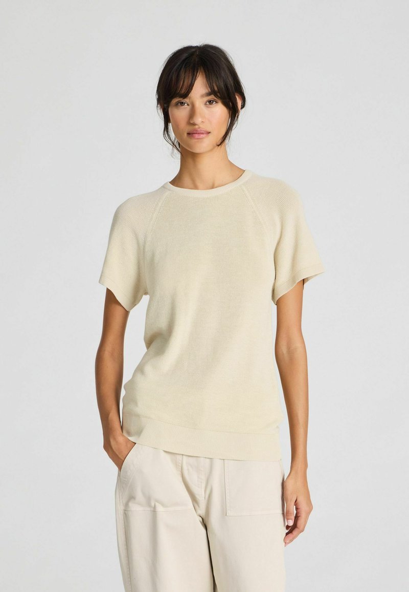 Light beige short-sleeve knit top with a round neckline and ribbed hem. Soft texture and relaxed fit, paired with cream-colored pants.