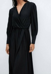 Robe noire à manches longues plissées, avec un décolleté en V profond et une ceinture en tissu à la taille, dotée d'une jupe fluide et d'un matériau texturé.