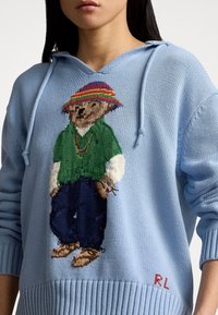 Light blue knitted hoodie featuring a bear graphic in green shirt and blue pants, with a multicolored striped hat and red embroidery.