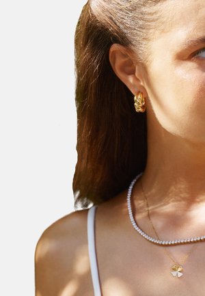 Gold hoop earrings with a twisted design, layered with a pearl necklace and a gold pendant featuring a floral motif. Straight brown hair visible.
