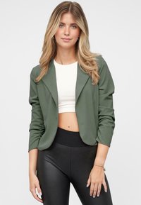 Blazer ligero de color verde oliva, con un corte entallado, frente abierto y mangas largas. Combinado sobre un top corto blanco y leggings negros.