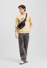 Sac bandoulière noir avec une finition lisse, doté d'une fermeture éclair et d'un cordon d'accent bleu. Porté avec un sweat-shirt beige et un jean gris.