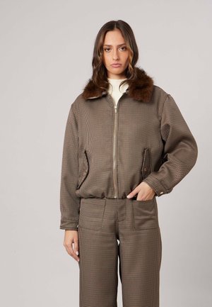 Vrouw draagt een bruin geruit jasje met bontkraag en bijpassende hoge taille broek, staand tegen een effen lichte achtergrond.