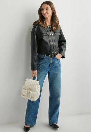 Kobieta w czarnej skórzanej kurtce, niebieskich dżinsach i czarnych butach, trzymająca biały pikowany plecak ze złotymi klamrami.