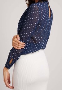 Blouse bleu marine transparente à pois blancs, manches longues, avec un détail en forme de goutte d'eau dans le dos. Caractérisée par une coupe ample et une fente ouverte aux poignets.