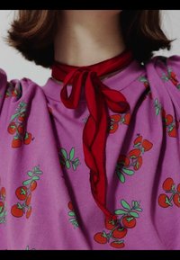 Personne portant une chemise violette avec un motif de tomates cerises rouges et un foulard rouge noué autour du cou, cadrée au niveau du menton et des épaules.