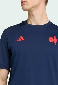 Mörkblåt tränings t-shirt med en röd Adidas-logotyp med tre ränder och ett rött höns-emblem på bröstet, tillverkad av slätt tyg.