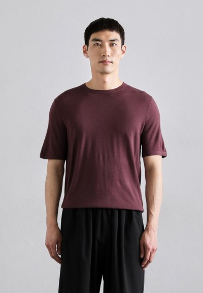 Bordeauxkleurig T-shirt met korte mouwen en ronde hals in een zachte, gestructureerde brei, gecombineerd met losse zwarte broeken, gedragen door een staande mannelijke figuur.