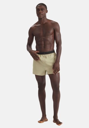 Ciemnoskóry, nagi mężczyzna stojący boso, ubrany w beżowe bokserki z czarną elastyczną gumką w pasie, na jednolitym jasnym tle.