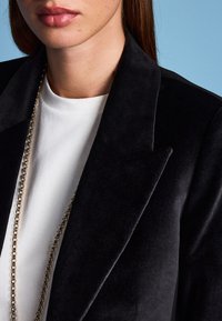 Blazer noir à texture veloutée, avec des revers aiguisés, porté sur une chemise blanche. Une délicate chaîne en or souligne l'encolure.