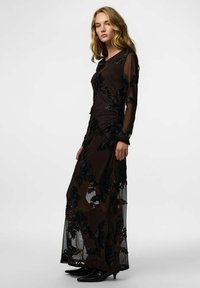 Robe longue noire en tissu transparent avec une broderie florale noire complexe. Elle présente des manches longues et une silhouette ajustée avec un léger éclat.