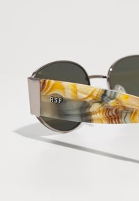Ochelari de soare rotunzi cu lentile întunecate; brațe din plastic marmorat galben, maro și gri; accente metalice; gravat "RSF" pe brațul stâng.