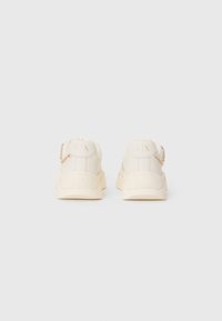 White sneakers with a textured surface, featuring a chunky sole, small logo on the heel, and a gold chain accent on the back.