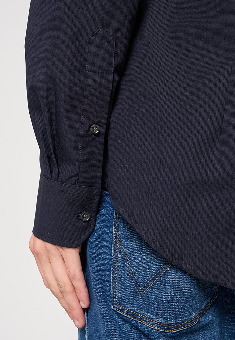 Dettaglio di una camicia blu navy con bottoni, caratterizzata da un polsino rotondo con due bottoni neri, abbinata a jeans in denim blu che mostrano un design distintivo della tasca.