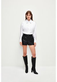 Camicia bianca con bottoni e maniche lunghe, pantaloni Corti di pelle nera, e stivali neri con tacco alti fino al ginocchio. Design semplice, vestibilità strutturata.