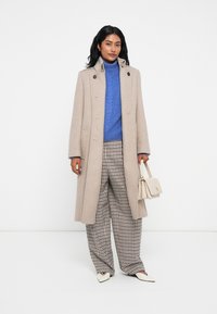 Cappotto di lana beige con un alto colletto, abbinato a un maglione blu a trecce, pantaloni a gamba larga marroni a quadro e una piccola borsa crema.