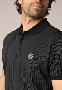 Polo shirt noir avec un motif texturé de petits points blancs, doté d'un col noir et d'un logo rond sur la poitrine gauche.