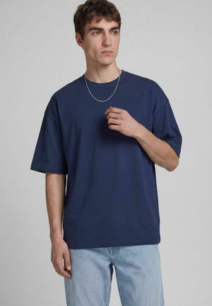 Jeune homme portant un T-shirt oversize bleu foncé et un jean bleu clair, debout devant un fond gris clair uni.