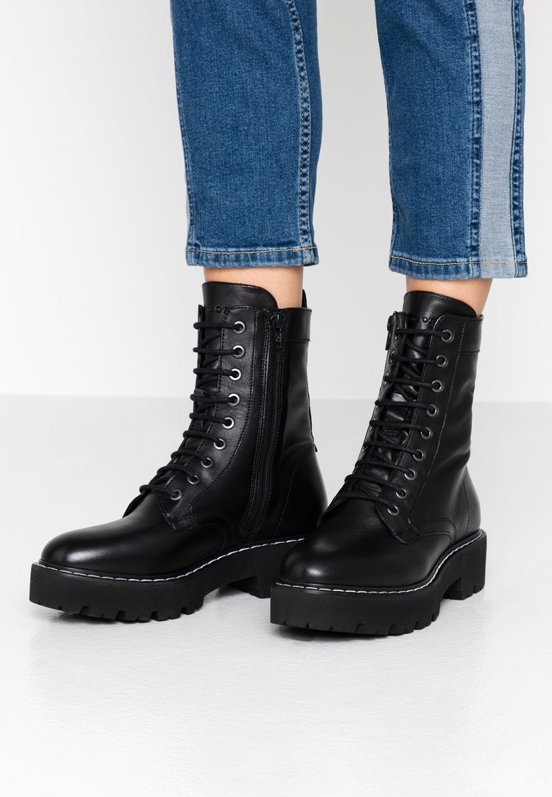 Bottes de combat en cuir noir à lacets avec fermetures éclair latérales, portées avec un jean en denim bleu coupé au-dessus de la cheville.