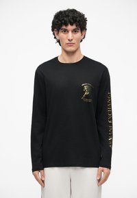 Garšs piedurkņu melns T-krekls, izgatavots no kokvilnas, ar zelta grafiku uz krūtīm un tekstu pa labo piedurknu. Vienkārša, taisna griezuma dizains.