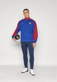 Kongelig blå jakke med røde ermer, med en logo, kombinert med marineblå ribbede bukser og hvite sneakers, holder en svart og gull fotball.