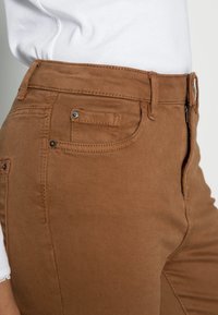 Hnědé bavlněné kalhoty s hladkou texturou, které mají postranní kapsu, páskové očka a zapínání na zip. Bílá košile je viditelná v oblasti pasu.