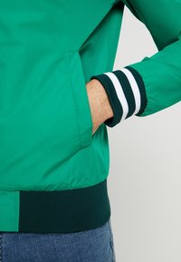 Grön bomberjacka med en slät yta, med vita och mörkgröna randiga ribbade ärmslut och sidofickor.