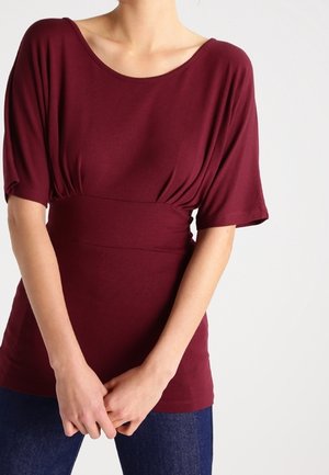 Bordeaux top met een wijde halslijn, korte mouwen en een ingezamelde taille, gemaakt van een zachte stof. Gecombineerd met donkerblauwe jeans.