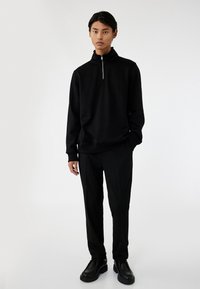 Zwarte pullover met een hoge kraag en een korte rits, gecombineerd met zwarte broeken en schoenen, tegen een neutrale achtergrond.