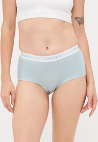 Egy nő világoskék hipster alsóneműt visel, fehér rugalmas derékpánttal, amelyen az "PIECES" felirat található, és egy fehér háttér előtt áll.