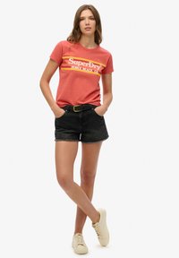 Camiseta de algodón roja con estampado en amarillo y naranja que dice "SuperDry Venice Beach, CA". Combinada con pantalones cortos de mezclilla negros y zapatillas color crema.