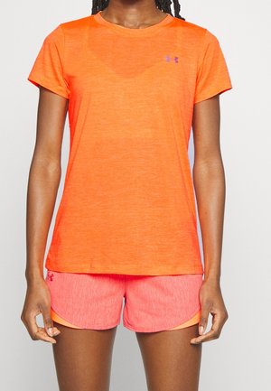 Person trägt ein leuchtend orangefarbenes kurzärmeliges Sportshirt und passende pink-orange Shorts und steht vor einem einfarbigen Hintergrund.