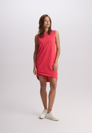 Bezramionna różowa sukienka o luźnym kroju i asymetrycznym dole. Wykonana z miękkiego, elastycznego materiału. Nosi ją z białymi trampkami, stojąc na neutralnym tle.