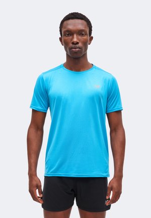 Hombre de pie mirando al frente, vistiendo una camiseta deportiva de manga corta azul brillante y pantalones cortos deportivos negros sobre un fondo claro y liso.